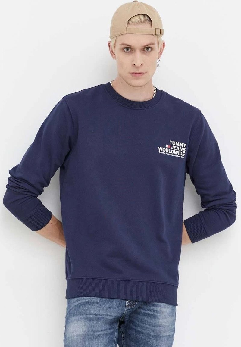 Felpa blu navale con polsini e orlo a coste, presenta un logo bianco sul petto. Indossata con jeans azzurri, su uno sfondo bianco.