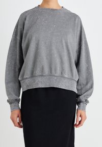 Persona vistiendo un sudadera gris desgastada y holgada de mangas largas, combinada con una falda negra, de pie frente a un fondo simple.