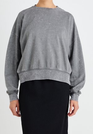 Persoon in een losse, vervaagde grijze sweatshirt met lange mouwen, gecombineerd met een zwarte rok, staande tegen een effen achtergrond.