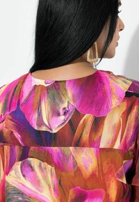 Kvinde med langt sort hår iført en lyserød og orange blomstret top samt stor geometrisk guldbøjle, set fra øvre ryg og sideprofil.