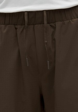 Brune bukser med elastik i taljen og snørre, båret med en hvid ribbet trøje, der viser talje- og øvre hofteområde.