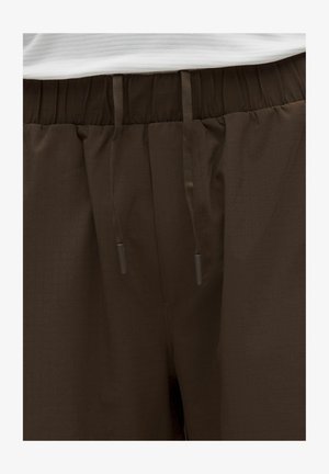 Brune bukser med elastik i taljen og snørre, båret med en hvid ribbet trøje, der viser talje- og øvre hofteområde.