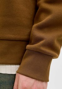 Brun sweatshirt stof med ribbede mansjetter. Glat tekstur, tætsiddende design, lagdelt over en lys stribet bomuldsbluse. Fokus på ærmedetalje.