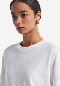 Hvid bomulds t-shirt med rund halsudskæring, der har en subtil tekstureret overflade. Modellen bærer guld hoop øreringe. Nærbillede fra skulderen.