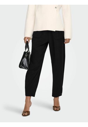 Femme portant un pantalon noir taille haute, blazer beige, sandales à talons marron, tenant un petit sac à main noir sur un fond clair.
