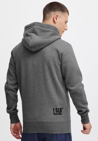 Grå hættetrøje med kengurulomme, ribbede manchetter og kant. Har logo-print på den nederste del af ryggen. Blødt stof, afslappet pasform.