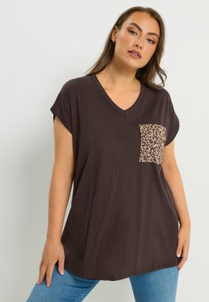 Kvinde iført en løs, mørkebrun V-hals top med en leopardmønstret brystlomme, kombineret med blå jeans, stående mod en ensartet lys baggrund.