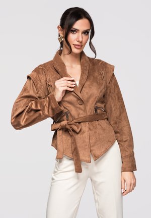 Kvinde iført en brun ruskindsjakke med bælte, lynlåslommer og skulderdetaljer, kombineret med hvide bukser mod en ensfarvet baggrund.