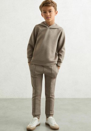Pojke som står med händerna i fickorna, iförd en taupe hoodie, matchande joggerbyxor och vita sneakers på grå golvbakgrund.