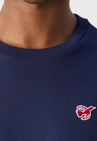 Marineblå bomulds T-shirt med rund halsudskæring og et lille rødt logo af en fugl broderet på brystet. Teksturen er glat og tætsiddende.