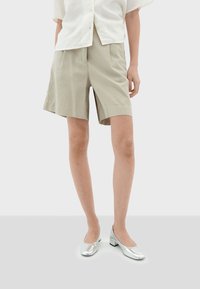 Beige shorts met een rechte snit, voorzien van plooien; gecombineerd met glanzende zilveren flats. De top is een losse, witte textuur shirt.