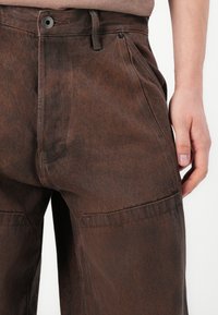 Primer plano de unos pantalones de mezclilla marrones con botón frontal, cremallera y bolsillos laterales; una mano reposa cerca del bolsillo derecho.