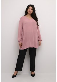 Rosa langärmlige Tunika mit V-Ausschnitt und lockerer Passform, kombiniert mit schwarzen geraden Hosen und spitzen schwarzen Absätzen. Glatte Textur.
