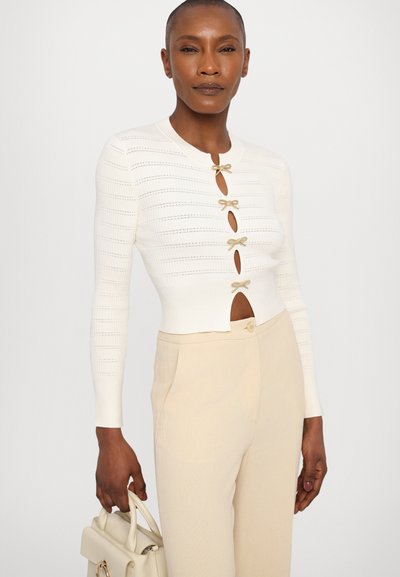 Hvit strikket cardigan med lange ermer, dekorert med gullsløyfe-detaller og nøkkelhullåpninger. Kombinert med beige skreddersydde bukser og en lys veske.
