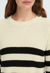 Maglione lavorato a maglia con una base crema e ampie righe orizzontali nere, un collo rotondo e una finitura testurizzata.