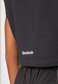 Juodas sportinis marškinėliai su lygiu paviršiumi, turintys baltą "Reebok" logotipą šalia apatinės siūlės. Marškiniai turi trumpas rankoves ir laisvą siluetą.