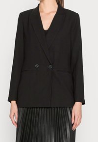 Kobieta ubrana w czarną marynarkę dwurzędową z dwoma guzikami, pod którą ma czarną bluzkę oraz czarną plisowaną spódnicę.