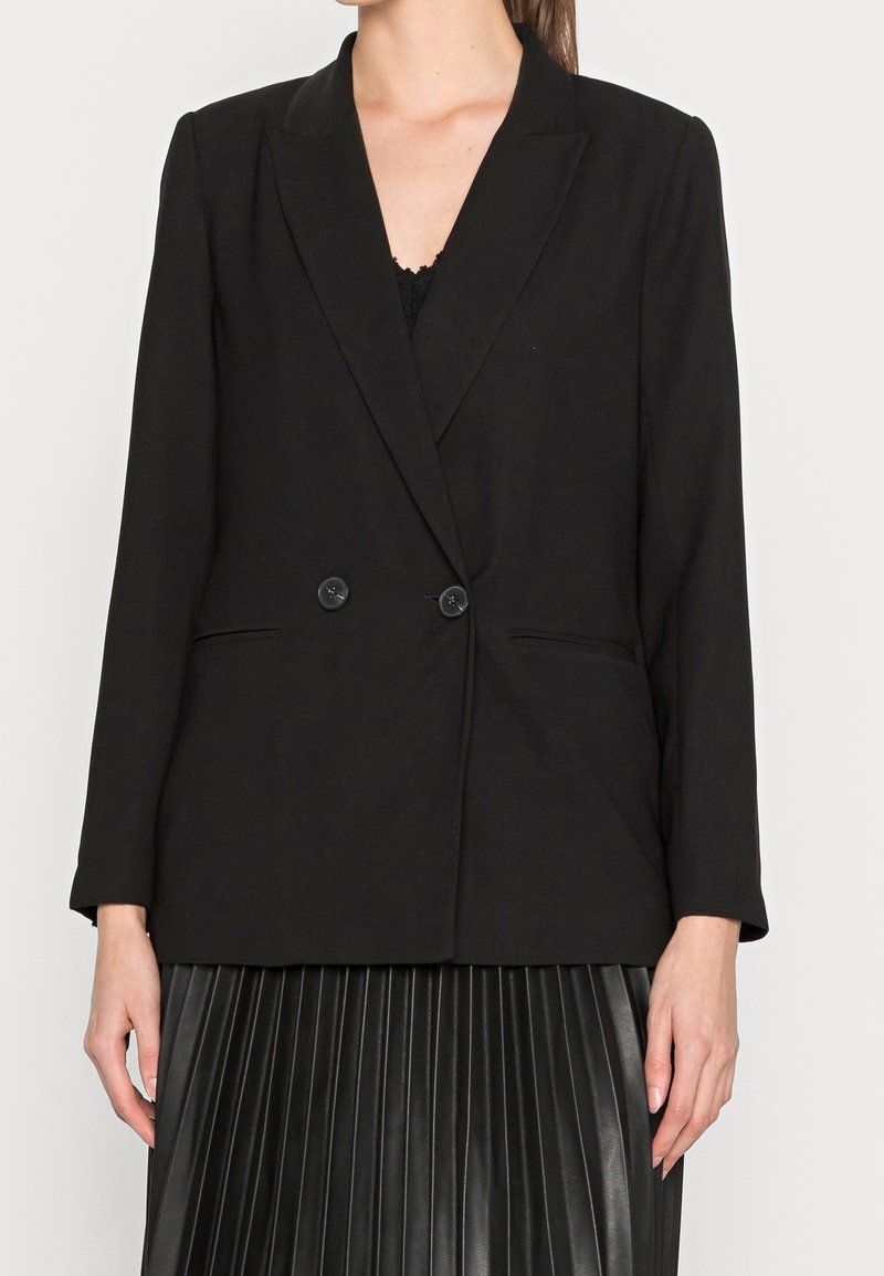 Kobieta ubrana w czarną marynarkę dwurzędową z dwoma guzikami, pod którą ma czarną bluzkę oraz czarną plisowaną spódnicę.