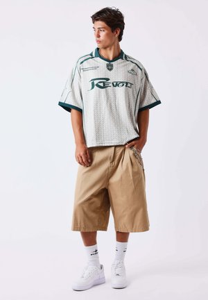 Jeune homme portant une chemise blanche et verte ample, un short beige au genou avec une chaîne, des chaussettes blanches et des baskets blanches, regardant sur sa gauche.