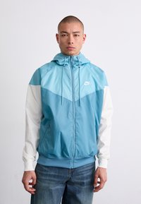 Gaiši zila vējjaka ar rāvējslēdzēju priekšpusē, ar baltām piedurknēm un kapuci. Iekļauj Nike logo uz krūtīm un sānu kabatām.