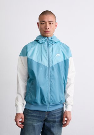 Coupe-vent bleu clair avec une fermeture éclair à l'avant, doté de manches blanches et d'une capuche. Comprend le logo Nike sur la poitrine et des poches latérales.