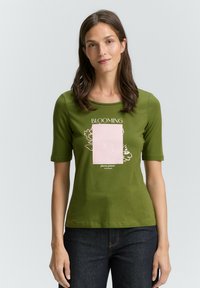 Groene katoenen T-shirt met korte mouwen en ronde hals, met een roze grafische rechthoek en bloemenontwerp. Donkere jeans maken de outfit compleet.