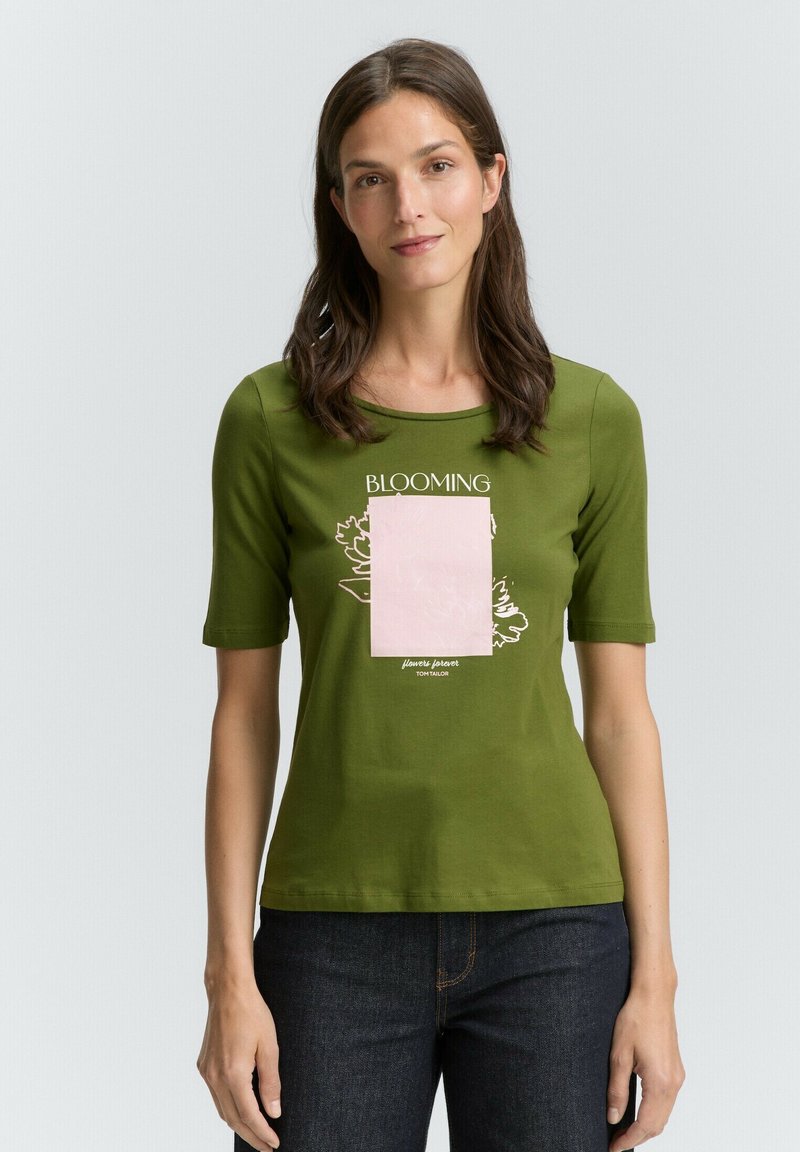 Groene katoenen T-shirt met korte mouwen en ronde hals, met een roze grafische rechthoek en bloemenontwerp. Donkere jeans maken de outfit compleet.