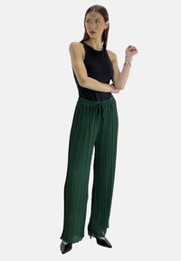 Zwarte mouwloze top gecombineerd met groene geplooide wijde broek met een elastische tailleband en trekkoord. Zwarte puntige schoenen.