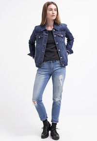 Denimjakke i mørkeblå med knappelukking, brystlommer og krage. Brukt over en svart topp og kombinert med lyse, slitte jeans og svarte støvler.