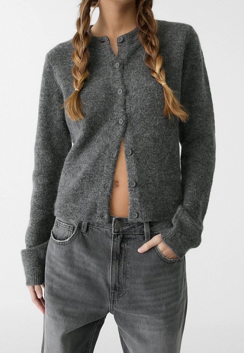 Personne avec deux tresses porte un cardigan gris boutonné et un jean en denim gris, la main glissée dans la poche avant, le ventre partiellement visible.