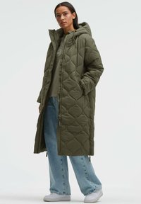 Olivgrüner, gesteppter langer Mantel mit Kapuze, ausgestattet mit Reißverschluss und Druckknöpfen, kombiniert mit hellblauen, weit geschnittenen Jeans und weißen Turnschuhen.