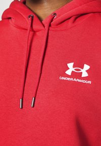Roter Hoodie aus weichem Stoff, mit einer Fronttasche, Kordeln mit metallischen Spitzen und einem weißen Under Armour-Logo auf der Brust.