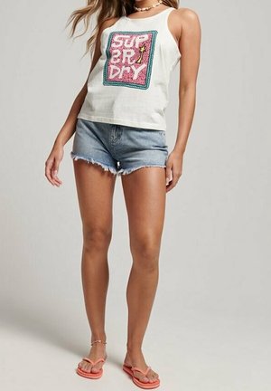 Woman wearing white sleeveless top with pink "Superdry" logo, denim cut-off shorts, and pink flip-flops standing against plain background.
