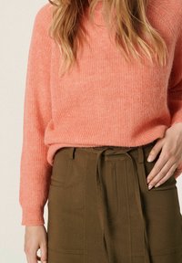 Pull rose texturé à manches longues, associé à une jupe marron avec une ceinture à nouer et des poches avant. Détails en tissu lisse visibles.