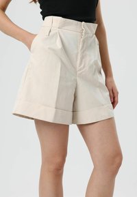 Beige op maat gemaakte korte broek met plooien bij de taille, ritssluiting en opgerolde boorden. De stof lijkt glad met een matte afwerking.