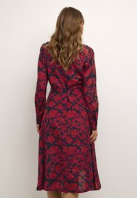 Donna con capelli ondulati che indossa un vestito lungo a maniche con una fantasia floreale rossa su sfondo blu, in piedi di spalle contro uno sfondo semplice.