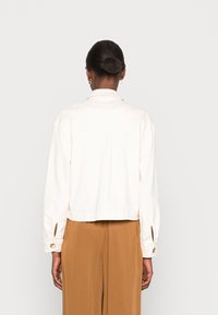 White, cropped jacket in a smooth fabric with a classic collar, buttoned cuffs, and rear seam detail, paired with brown wide-leg pants.