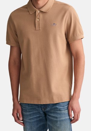 Homme portant un polo beige à manches courtes avec deux boutons et un jean bleu, debout face à l'objectif, haut du corps visible.