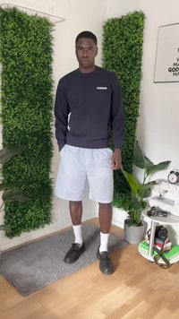 Vyras stovi apsirengęs juodu sportiniu megztiniu, juostuotais šortais, baltais kojiniais ir juodais loaferiais kambaryje su augalais ir fotografijos įranga.