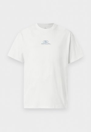 Jeune homme aux cheveux courts et bouclés, portant un t-shirt blanc à manches courtes avec un petit logo et un pantalon cargo beige, debout devant un fond uni.