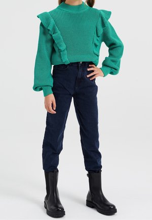 Enfant portant un pull en maille turquoise à volants, un pantalon taille haute bleu foncé, et des bottines noires, debout avec une main sur la hanche.