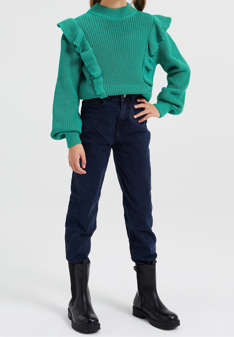 Enfant portant un pull en maille turquoise à volants, un pantalon taille haute bleu foncé, et des bottines noires, debout avec une main sur la hanche.