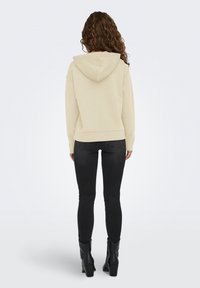 Donna dai capelli ricci che indossa un hoodie beige, jeans skinny neri e stivaletti neri con tacco, in piedi di spalle su uno sfondo neutro.