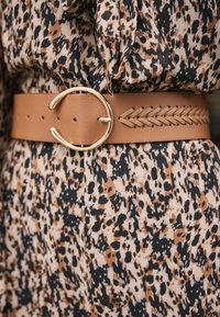 Läderbälte i tan med en cirkulär guldbuckle och en flätad design längs kanten, bärs över en klänning i svart och leopardmönster.