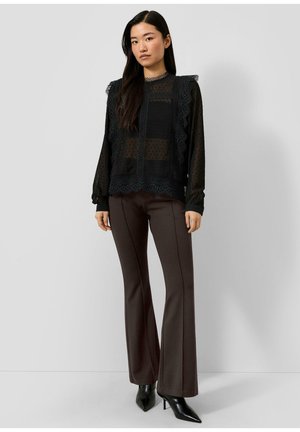Vrouw die een zwarte kanten blouse met doorschijnende mouwen draagt, donkerbruine uitlopende broek en zwarte puntige hoge hakken, staand tegen een effen achtergrond.