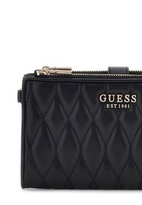 Portafoglio in ecopelle trapuntata nera con zip e accessori dorati. Presenta il logo "GUESS EST 1981" impresso sul fronte. Design rettangolare compatto.