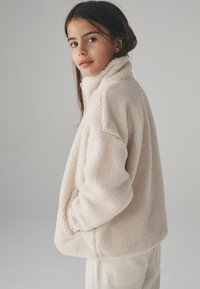 Mjuk beige fleecjacka med hög krage, sidofickor och avslappnad passform, med en texturerad yta och enkelt mönster.