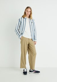 Gestreiftes Button-Down-Hemd in Blau und Weiß, kombiniert mit einem weißen T-Shirt, beigen weit geschnittenen Hosen und navyblauen Turnschuhen mit grauen Akzenten.