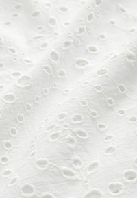 Tessuto bianco con intricate lavorazioni ricamate, caratterizzato da ritagli circolari e disegni floreali, che mette in evidenza una superficie texturizzata.