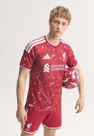Ung mand i rød Liverpool FC fodboldtrøje holder bold i samme farve mod ensfarvet baggrund.